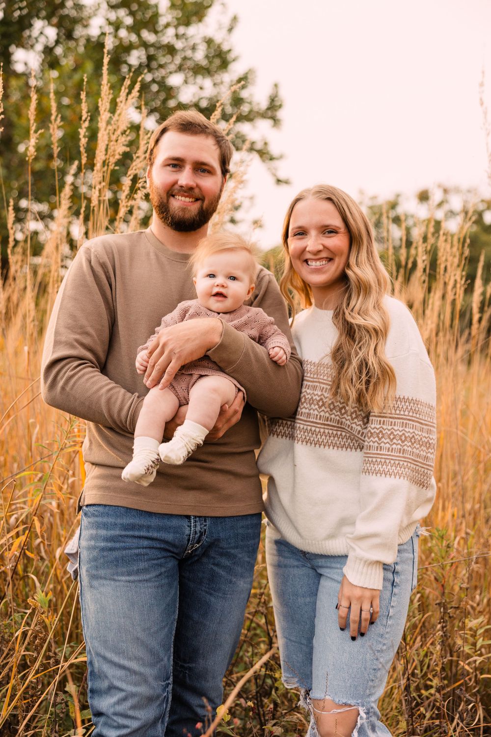 Smiling couple holds their baby in a golden field of tall grass, wearing cozy sweaters and jeans for a family portrait.
