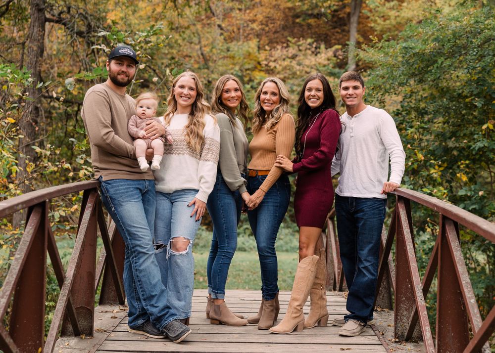 Family of seven with a baby poses on a wooden footbridge surrounded by colorful autumn trees in a fall portrait.