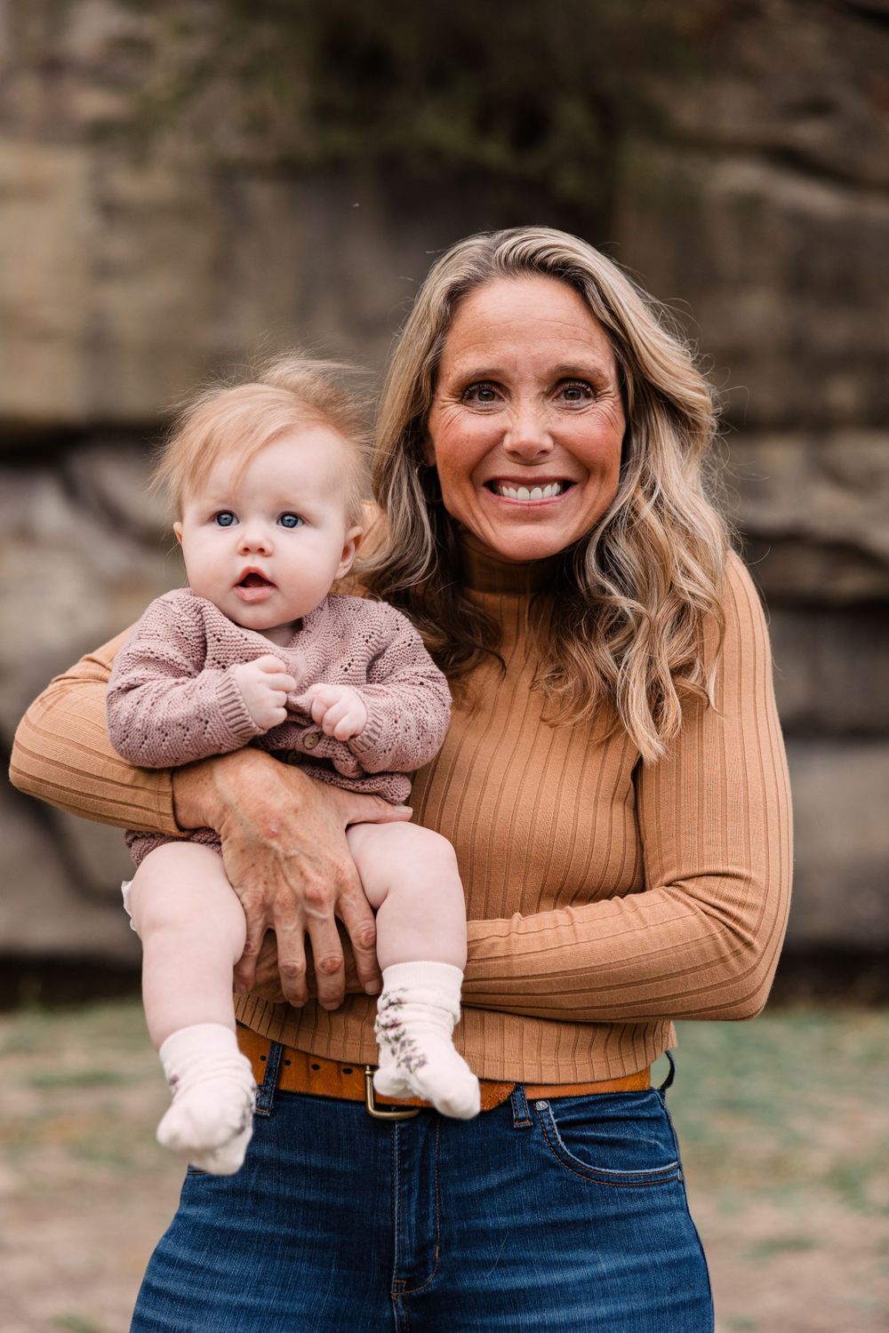Smiling woman with wavy blonde hair holds a baby in a pink knit sweater in front of a rocky bluff.