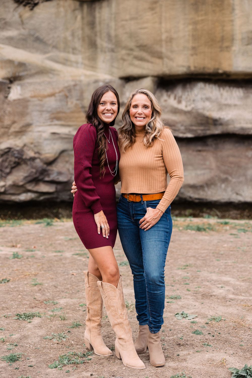 Two smiling women in fall outfits and boots pose arm in arm in front of a natural rock wall.