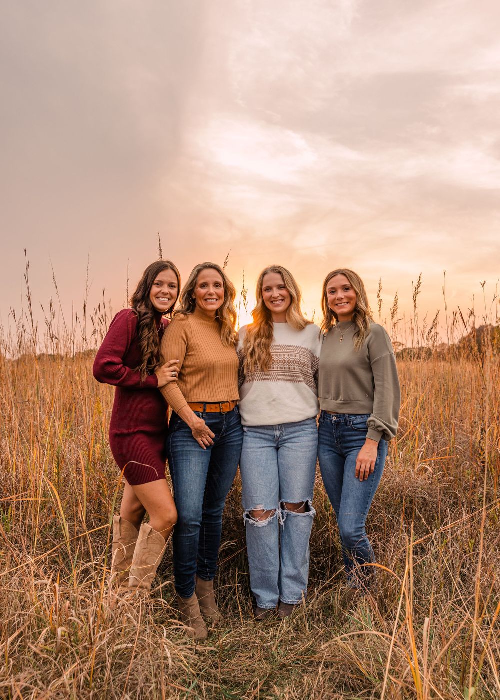 Four smiling women in cozy sweaters and jeans pose together in a tall golden grass field at sunset.