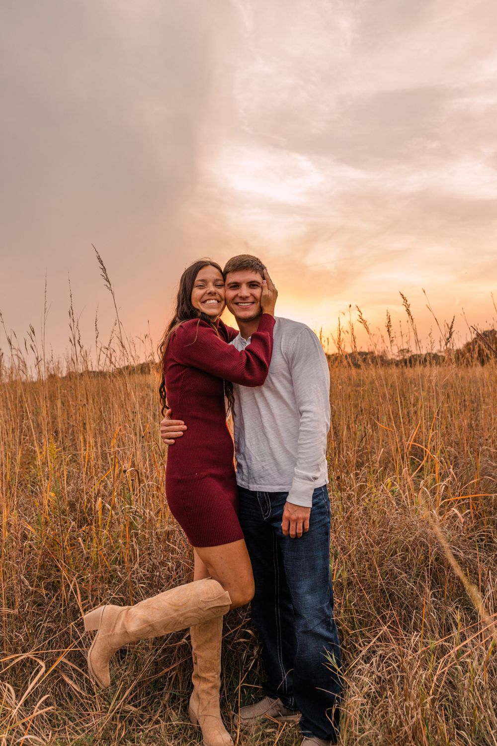 A smiling couple embraces in a golden prairie at sunset, she wearing a burgundy dress and tan boots.