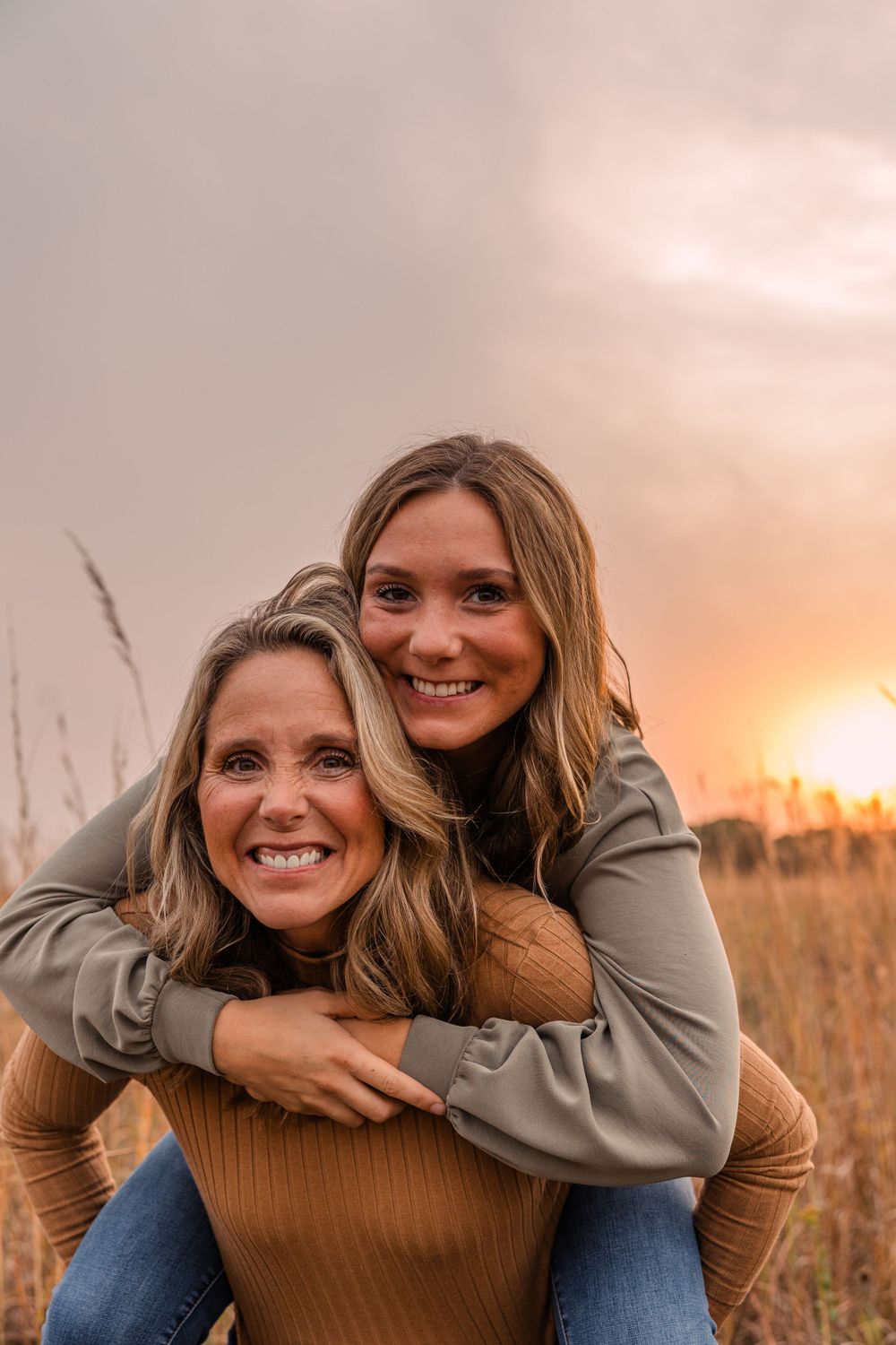 Two smiling women laugh together as one gives the other a piggyback ride in a field at golden hour.