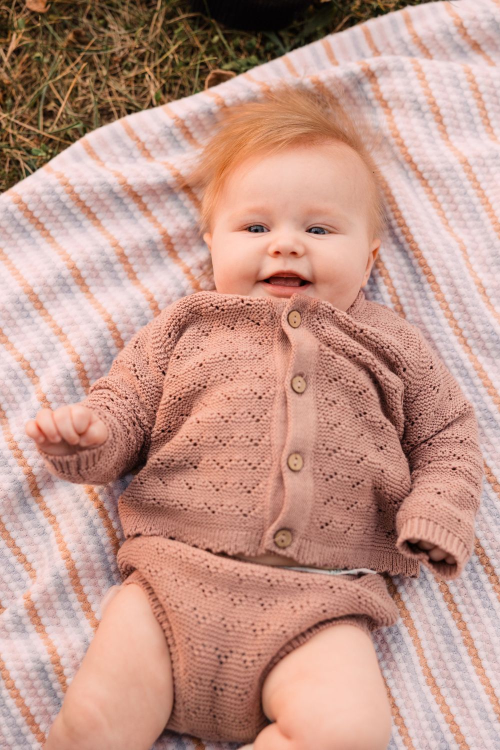 A smiling baby with strawberry blonde hair lies on a striped blanket wearing a dusty pink knit cardigan and bloomers.
