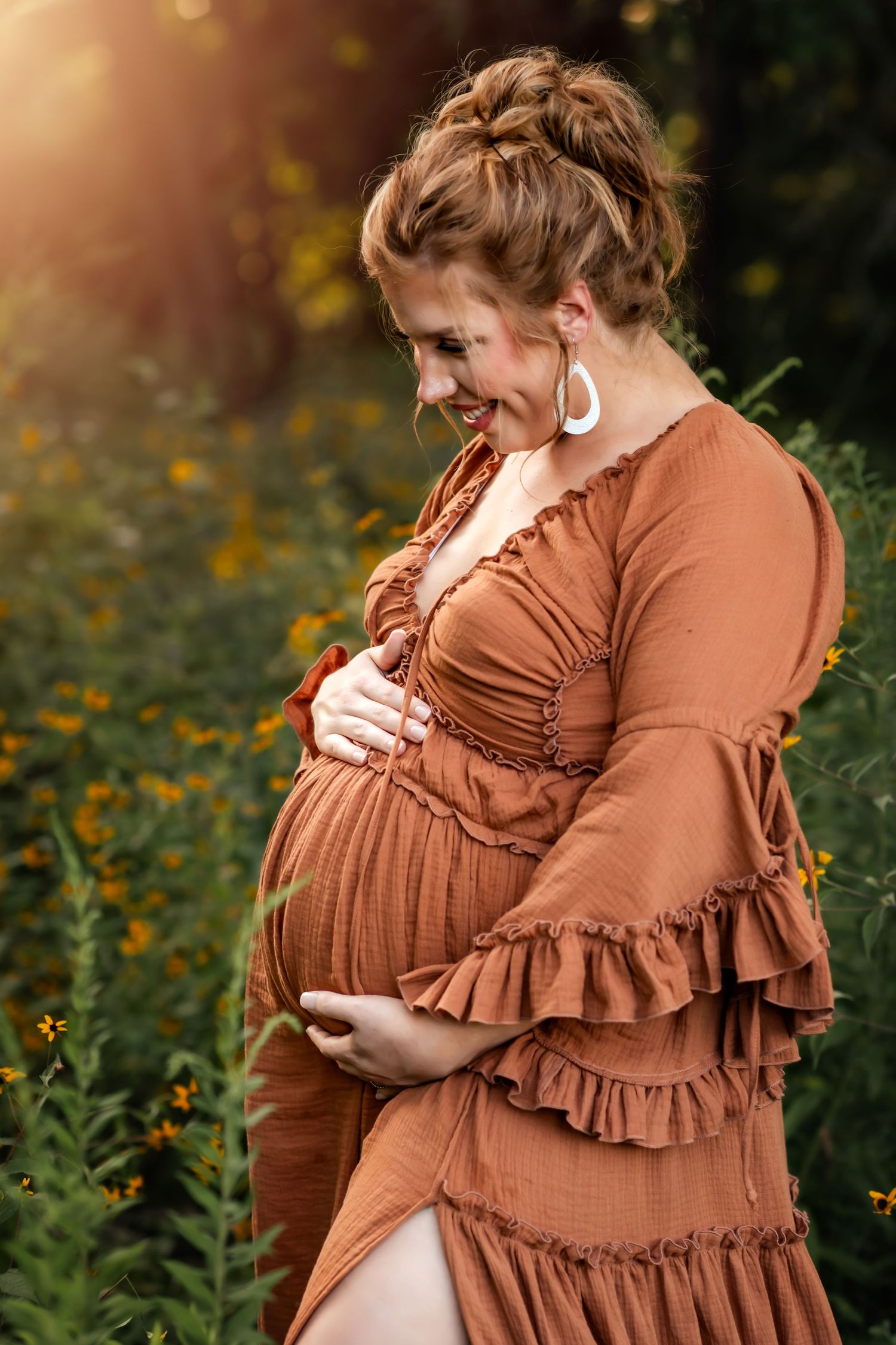 A maternity portrait in a rust-colored dress amid wildflowers at sunset.