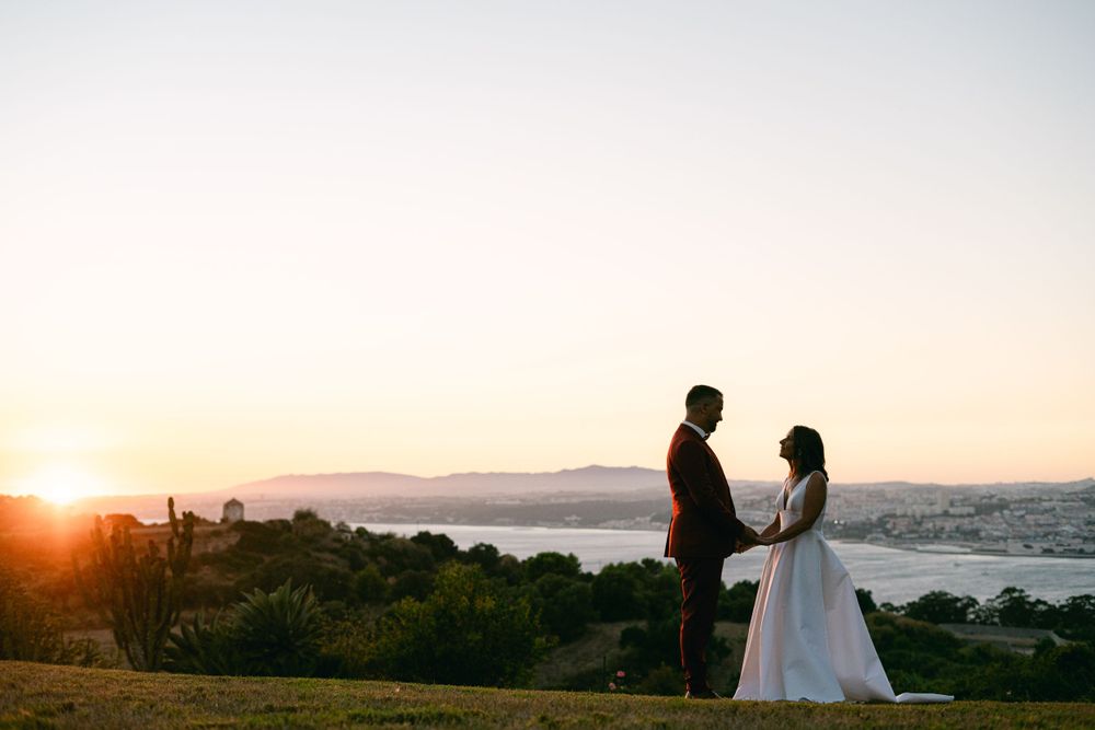 A couple stands in silhouette at sunset overlooking a scenic mountain vista and bay during an outdoor wedding ceremony.