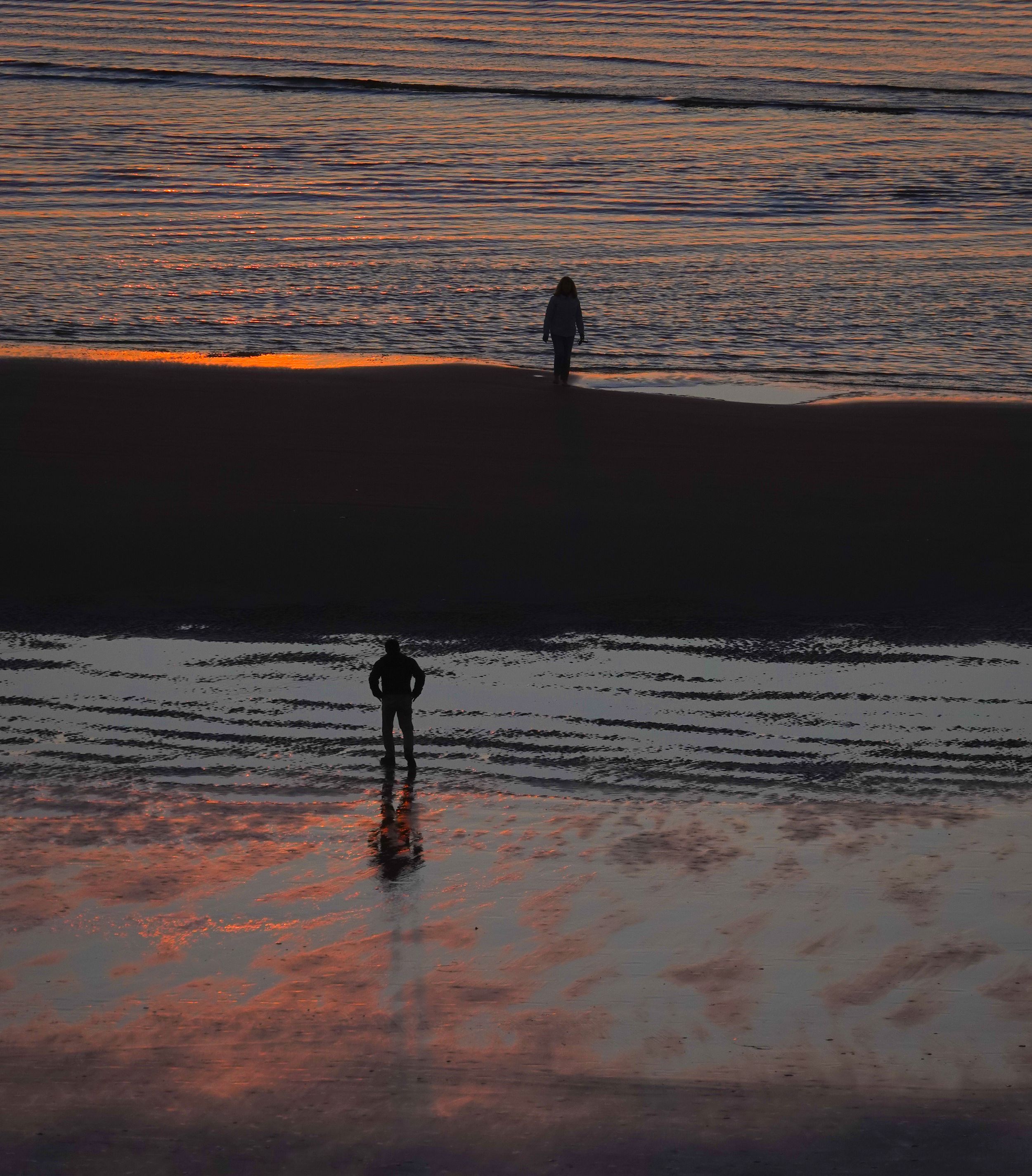 Silhouetted figures stand on beach at sunset with orange reflections in wet sand.