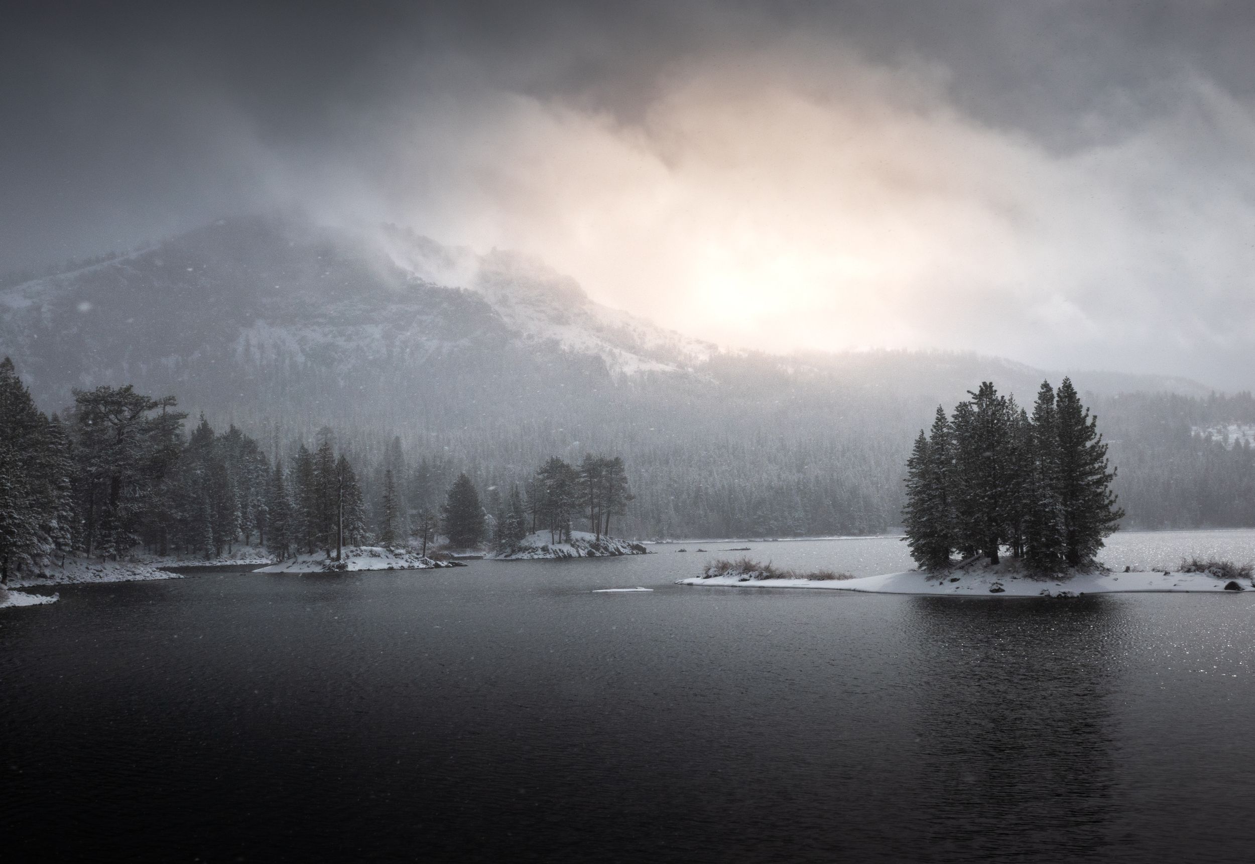 Misty winter lake scene with snow-covered mountains and evergreen trees reflected in dark waters.
