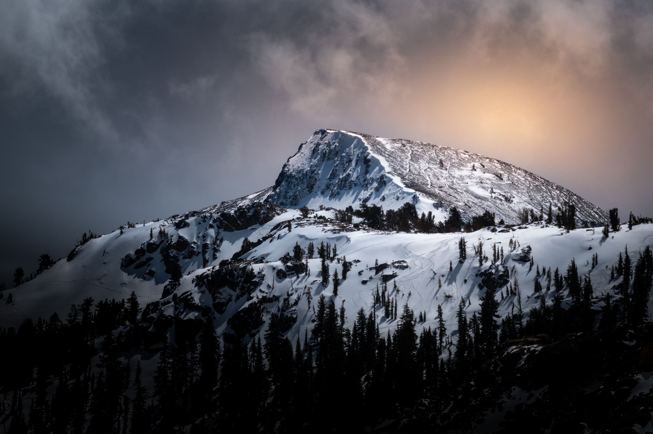 Snow-covered mountain peak glows under dramatic stormy sunset sky.