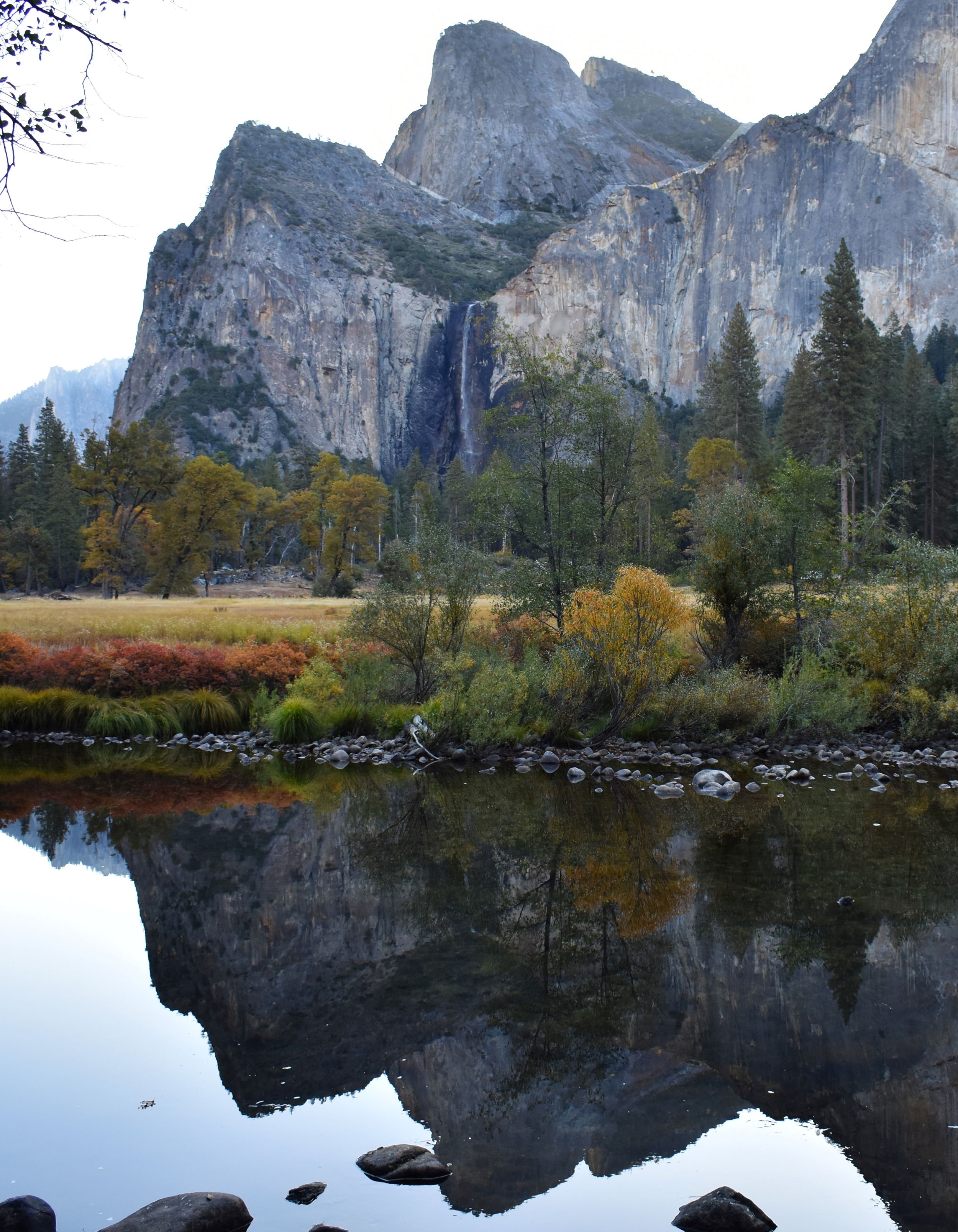 Dramatic granite cliffs of Yosemite Valley reflect in calm waters with autumn foliage along the shore.