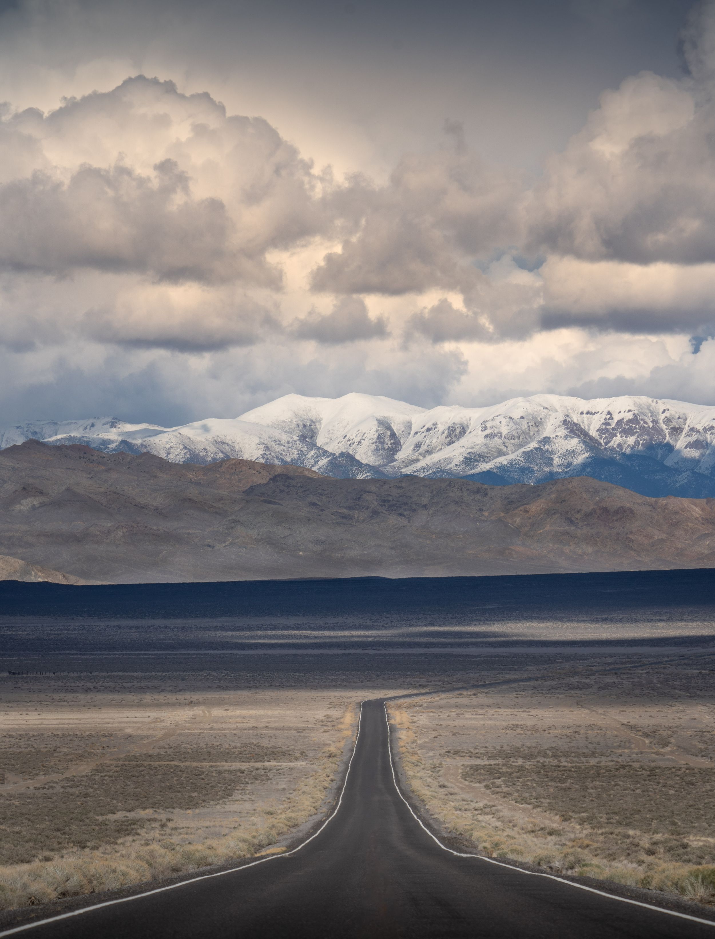 A dramatic desert highway stretches toward snow-capped mountains under stormy clouds in a vast open landscape.