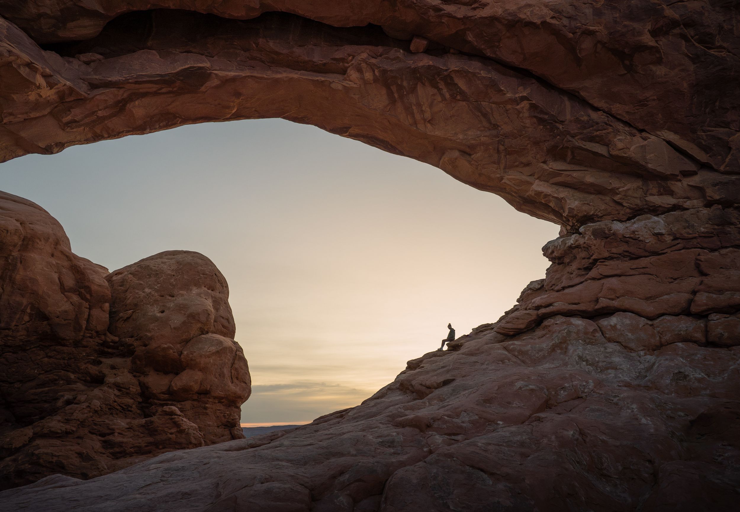 Natural stone arch formation in red sandstone desert landscape silhouetted against a bright evening sky.