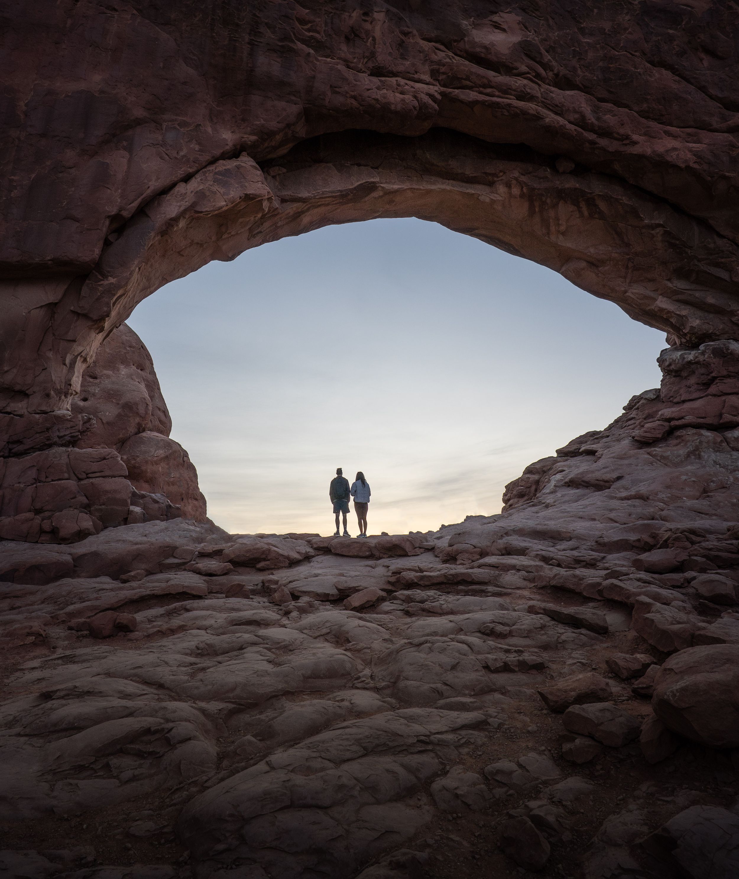 Two silhouettes stand framed within a natural rock arch formation at sunset in a desert landscape.