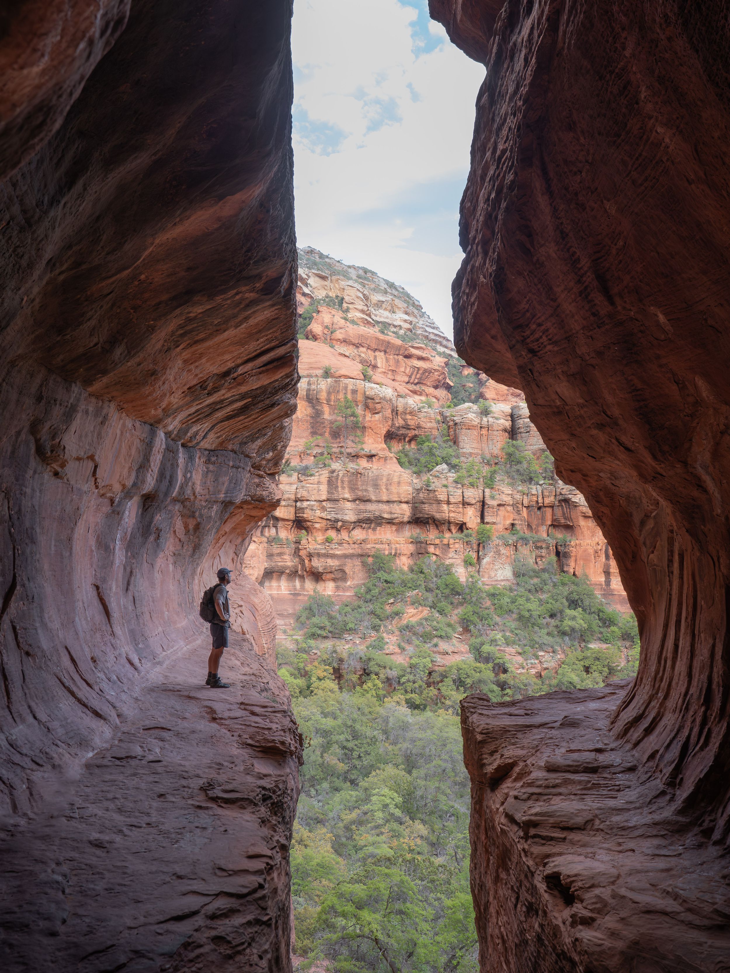 A silhouetted hiker stands in a natural cave opening framed by red rock formations in a canyon landscape.