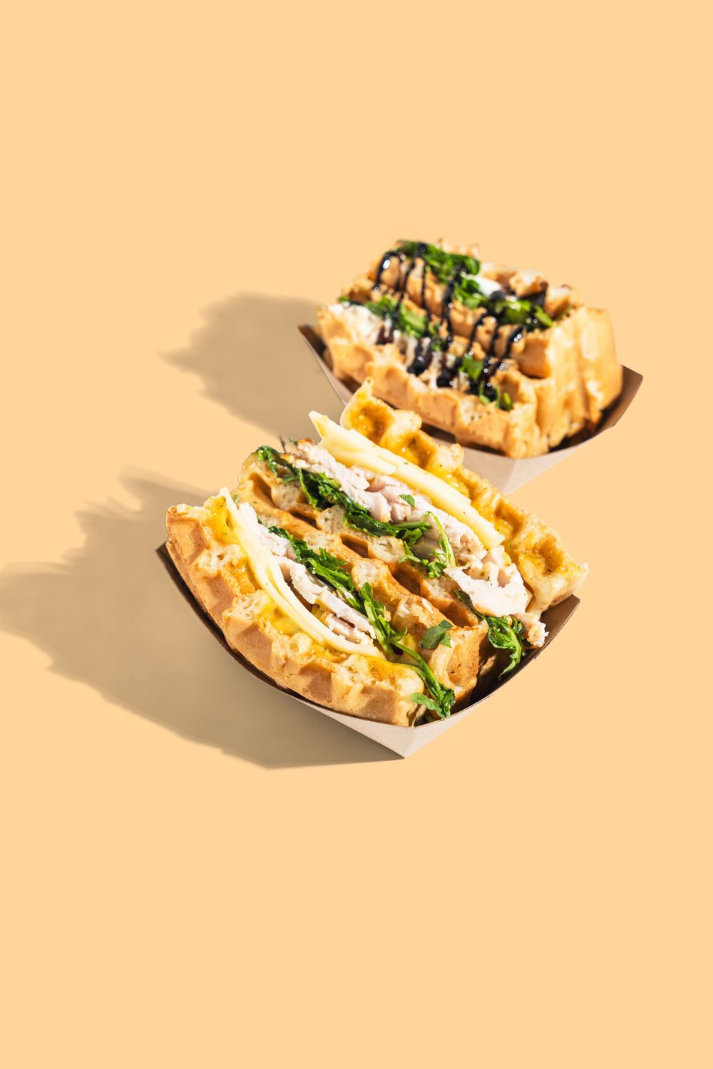 Two loaded waffle dishes with sauce and herbs in paper trays on peach background. Mtlshutter photography