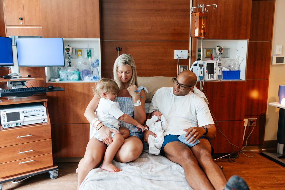 Family sitting together on hospital bed with medical equipment and wooden paneled walls in the background.