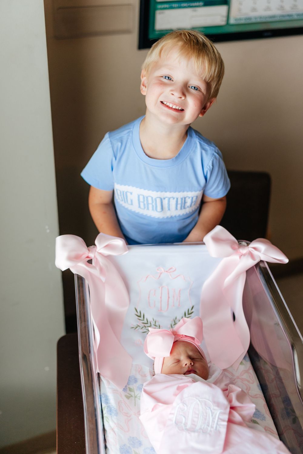 Young child in light blue shirt smiles while looking at newborn baby in pink blanket and bow.
