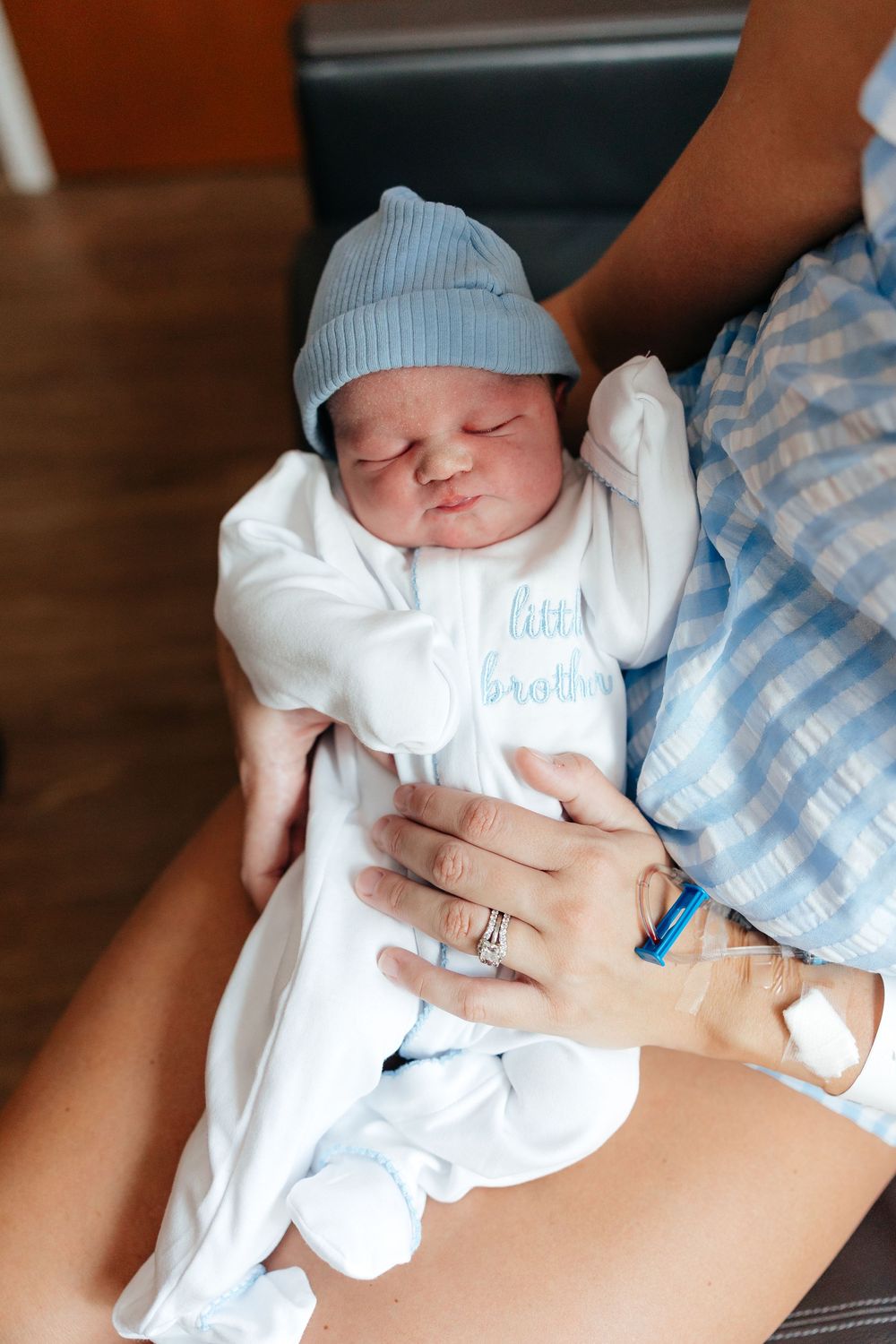 Sleeping newborn baby wearing blue knit hat and white onesie rests in parent's arms with engagement ring visible.