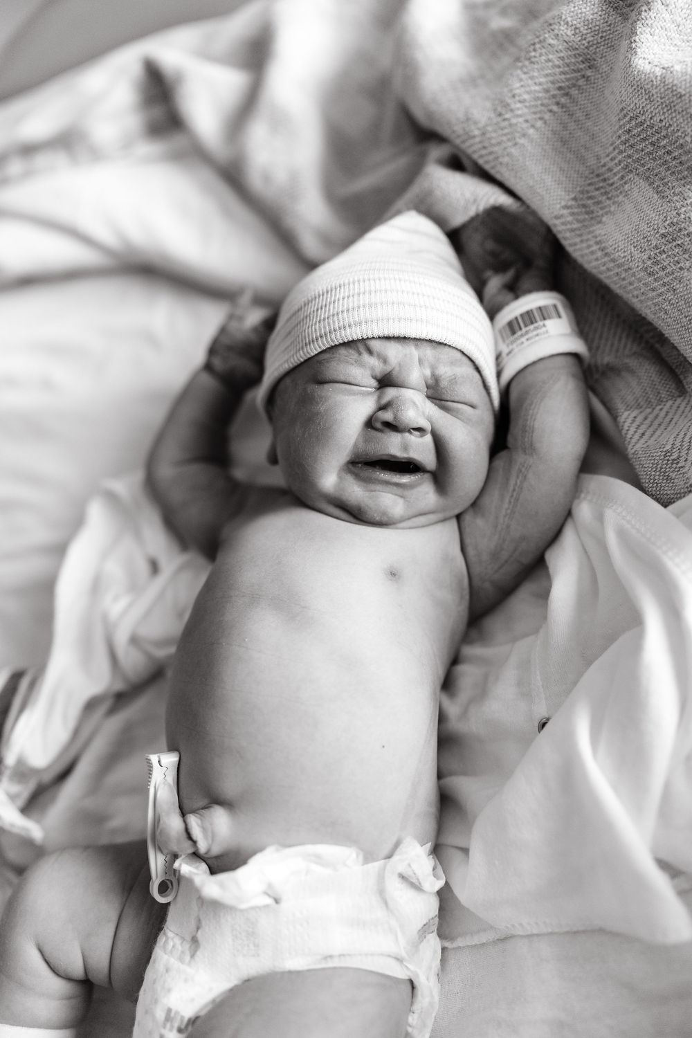 A newborn baby in a white hat cries while lying on white hospital bedding in black and white photography.