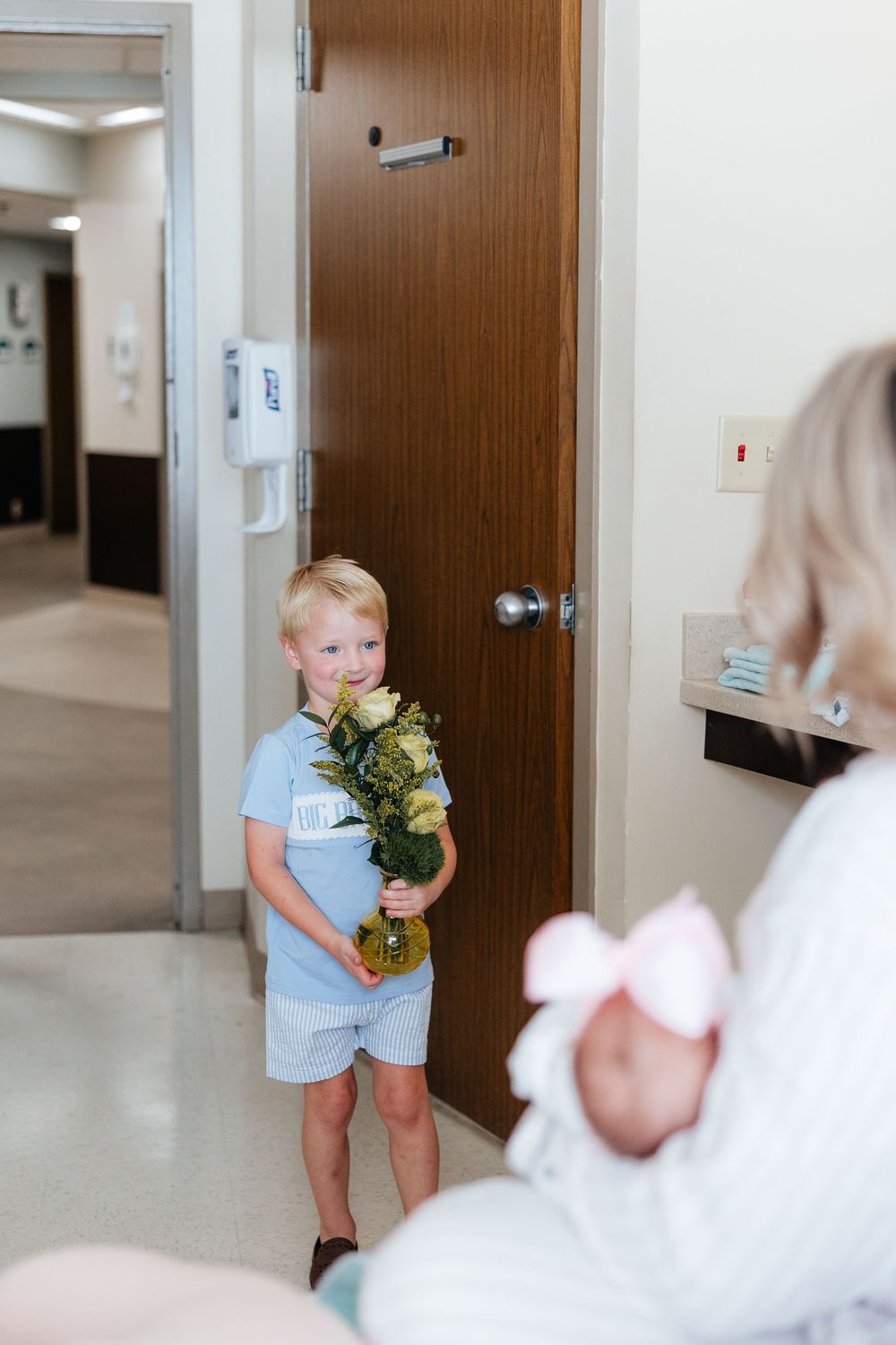 A small child in light blue clothing carries a bouquet of flowers down a hallway with wooden doors.