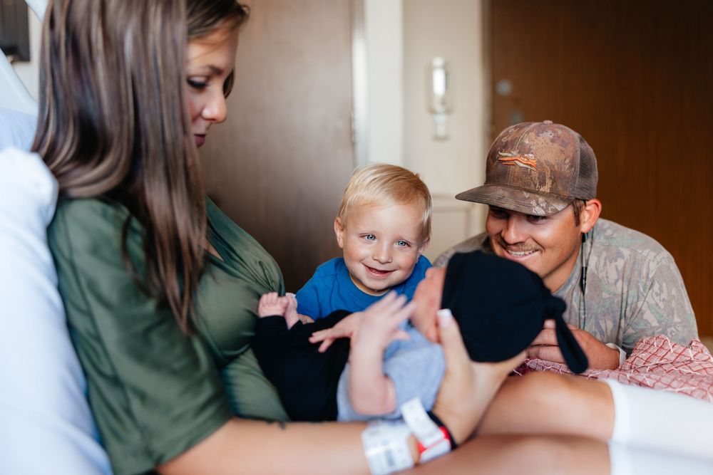 A young family smiles together in a hospital room, with a toddler boy in a blue shirt reaching out happily.