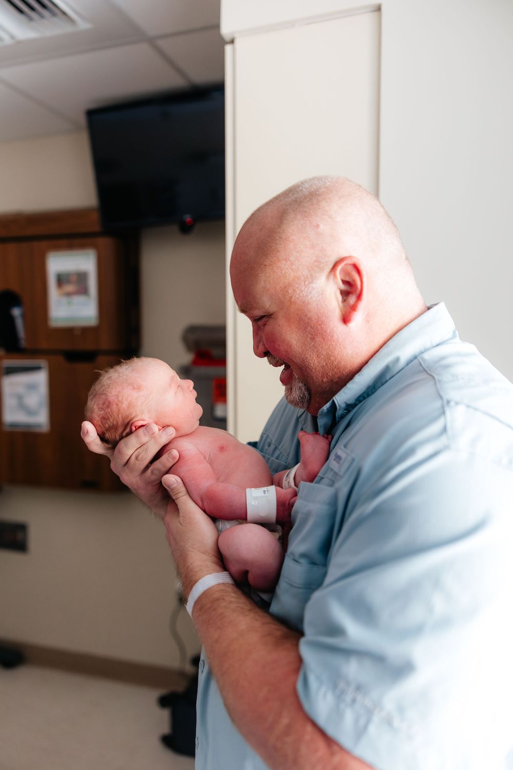Bald father tenderly holds newborn baby in hospital room, gazing lovingly at his child after birth.