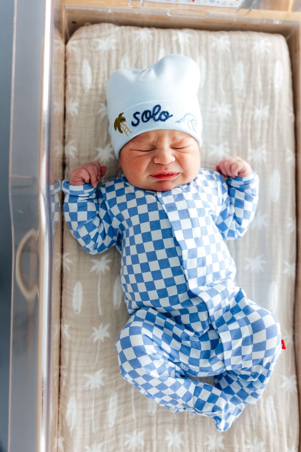 Baby wearing blue and white checkered sleepwear rests peacefully on white bedding.