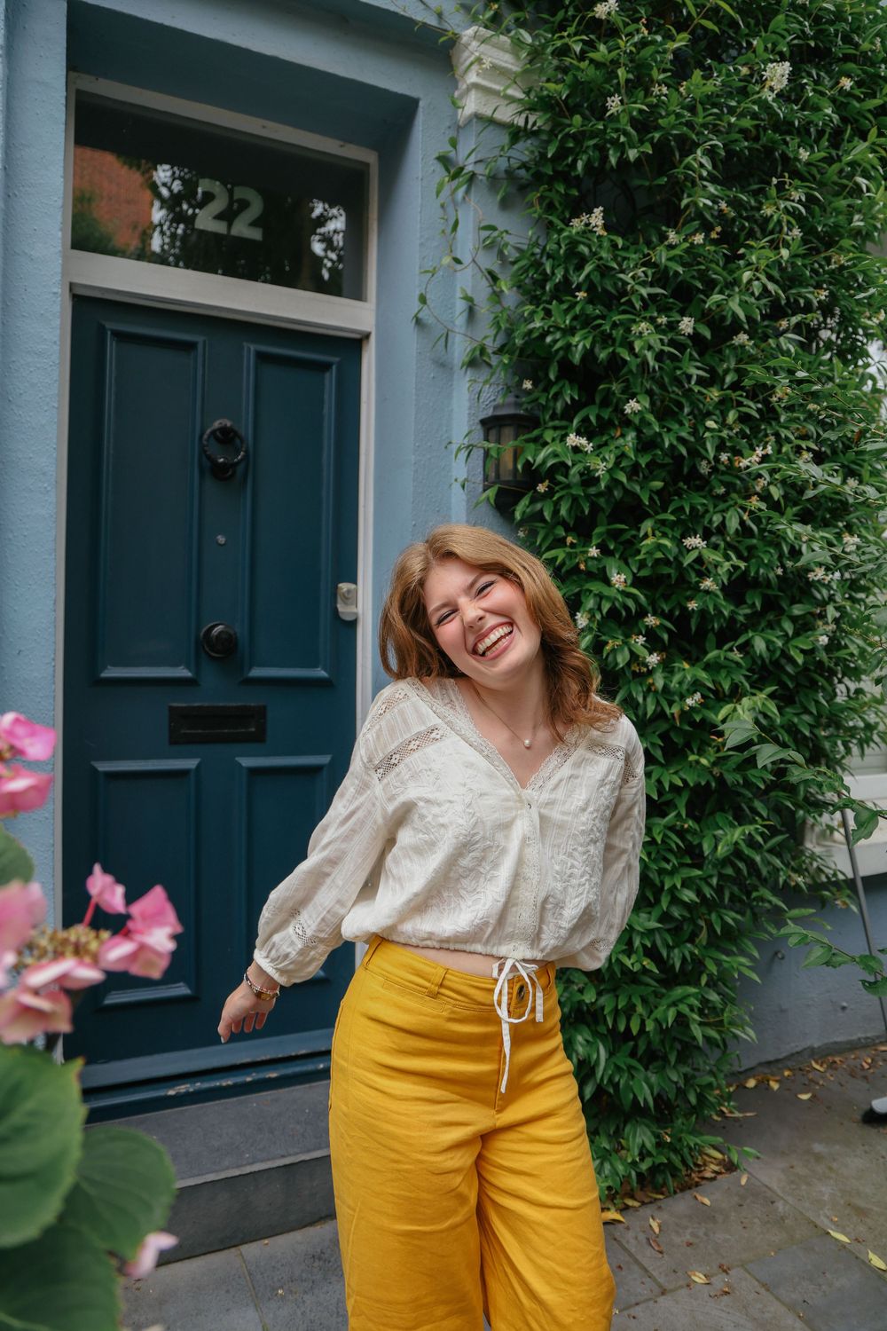Smiling young woman in yellow trousers and white blouse standing outside a house with teal door.
