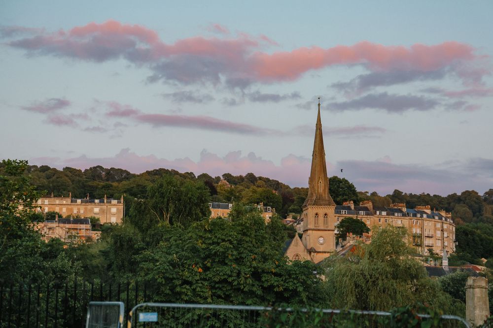 A church spire rises above trees and Georgian buildings at sunset with pink clouds in the sky.