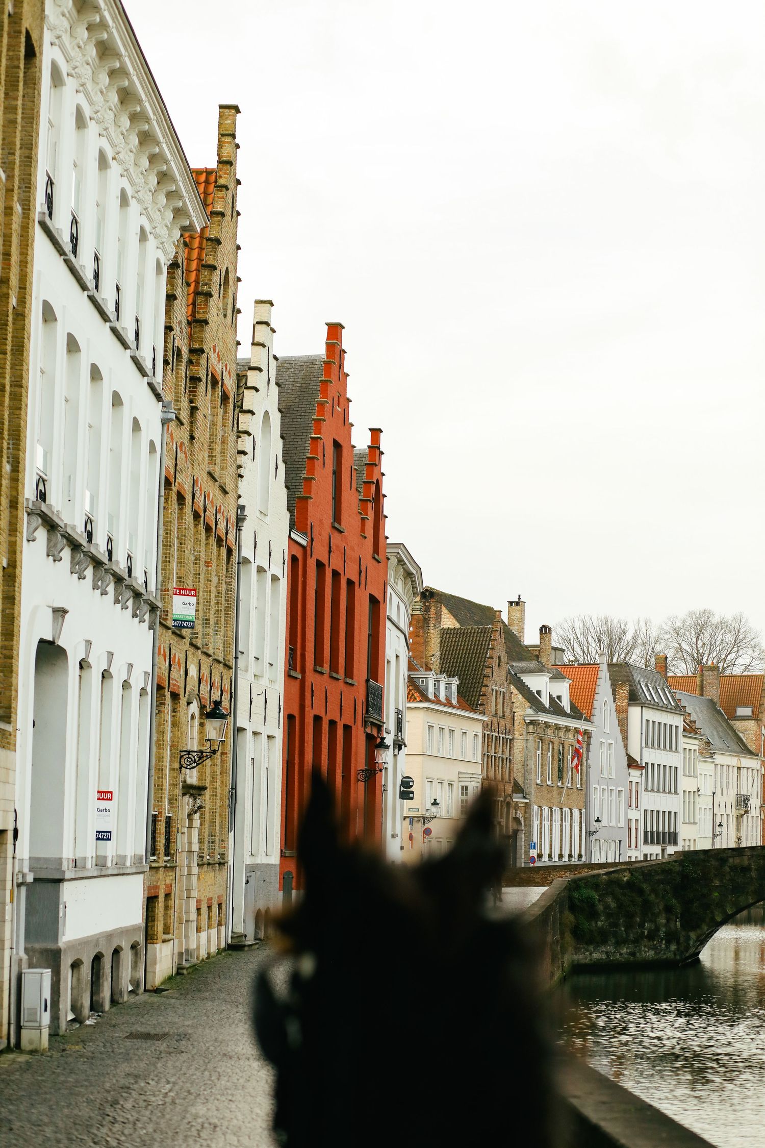 Colorful historic stepped-gable buildings line a canal street in Bruges, Belgium on an overcast day.