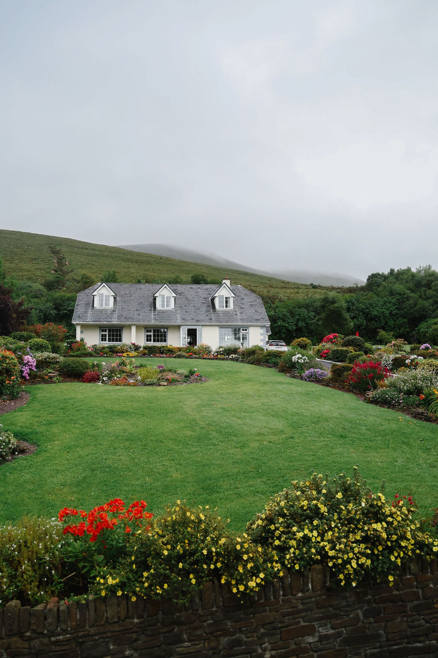 A charming white Irish cottage surrounded by colorful flower gardens with rolling green hills in the background.