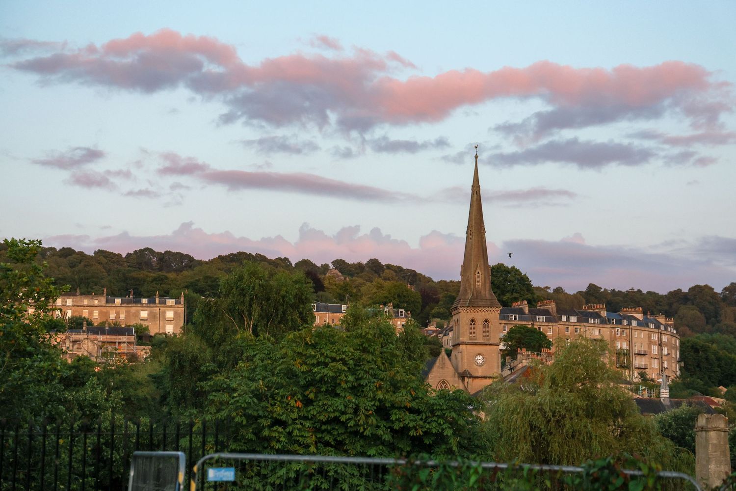 A historic church spire rises above lush trees at sunset with pink clouds in a small European town.
