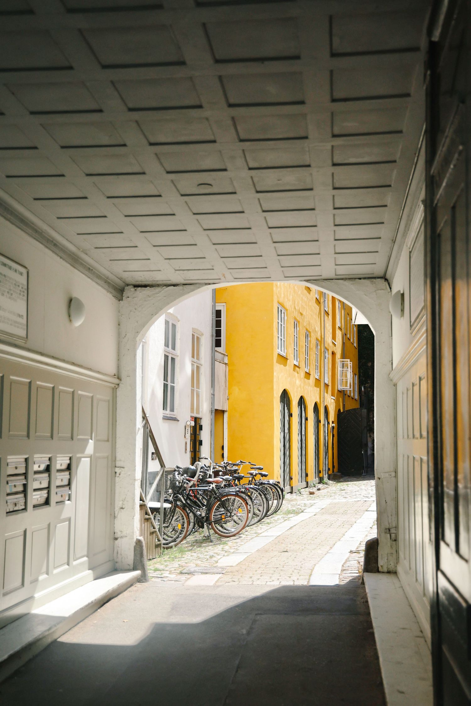 A sunlit cobblestone courtyard with bicycles parked beside a vibrant yellow building viewed through an archway.