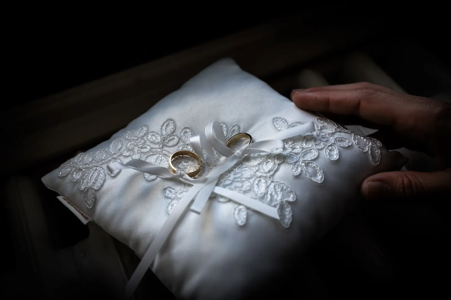 Deux alliances posées sur un coussin de satin blanc brodé, tenues délicatement par une main dans une lumière tamisée.