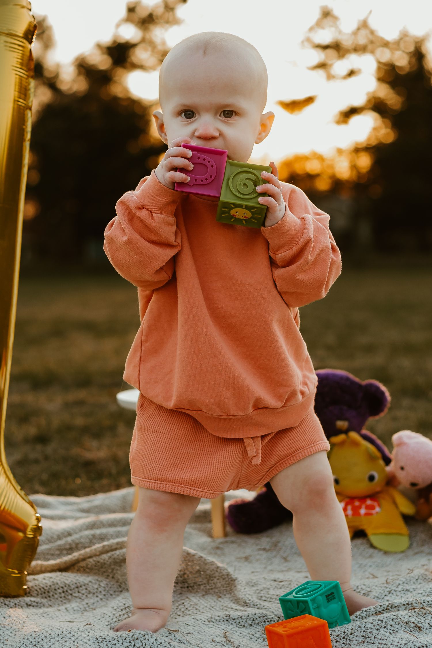 Baby in orange outfit lies on blanket next to large gold number one balloon during sunset photoshoot outdoors.