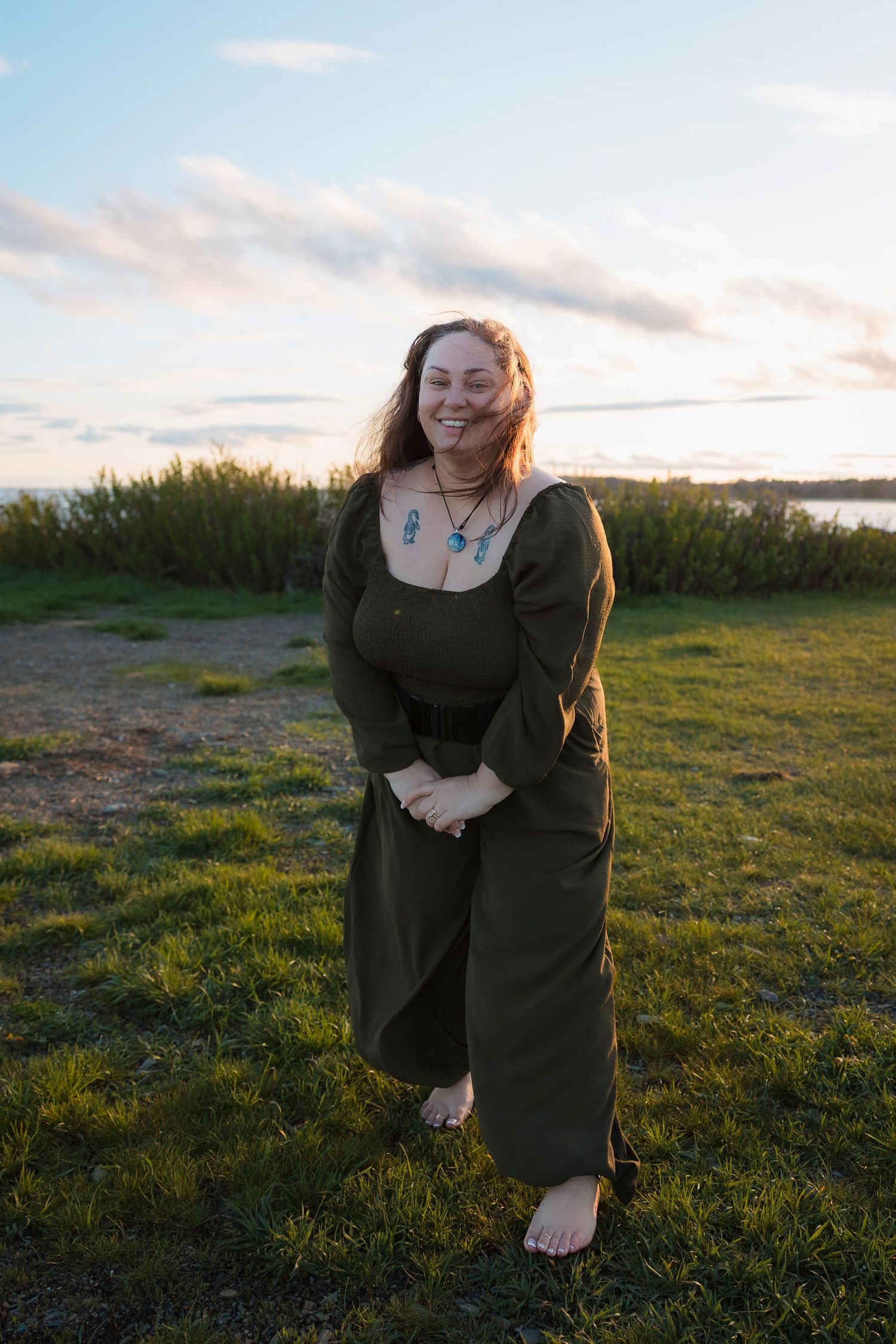 A person wearing a dark green jumpsuit stands barefoot on grass at sunset near a waterfront.