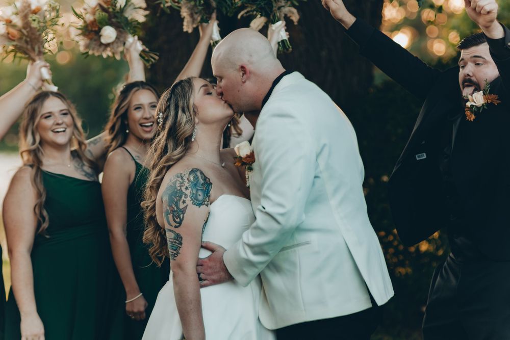 A romantic kiss at an outdoor wedding with guests watching and smiling in the background.