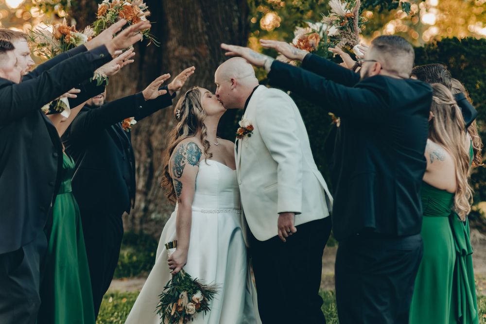 Wedding couple shares a kiss under an archway of bridesmaids in green dresses and groomsmen during outdoor ceremony.