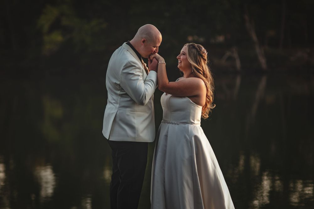 Emotional wedding couple shares a tender sunset moment by a lake in formal white attire.