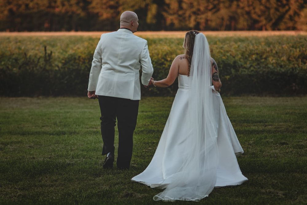 A bride in a flowing white dress and veil walks through a grassy field at sunset with a companion in a light gray suit.