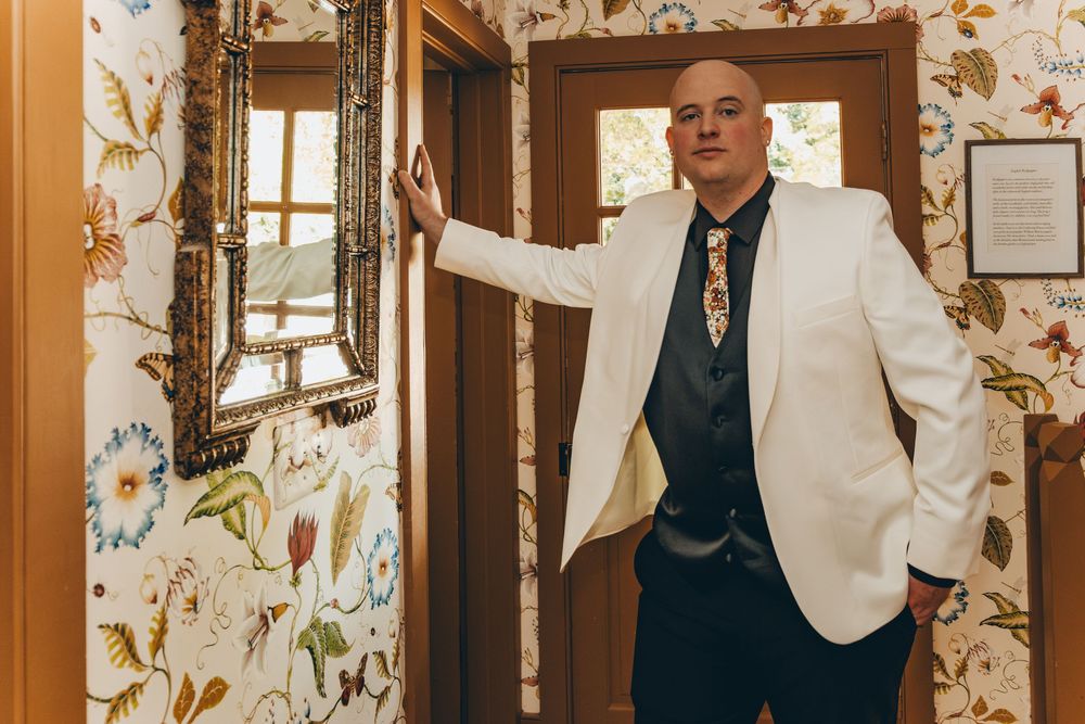 A person wearing a white blazer and dark tie stands confidently in a doorway with floral wallpaper.