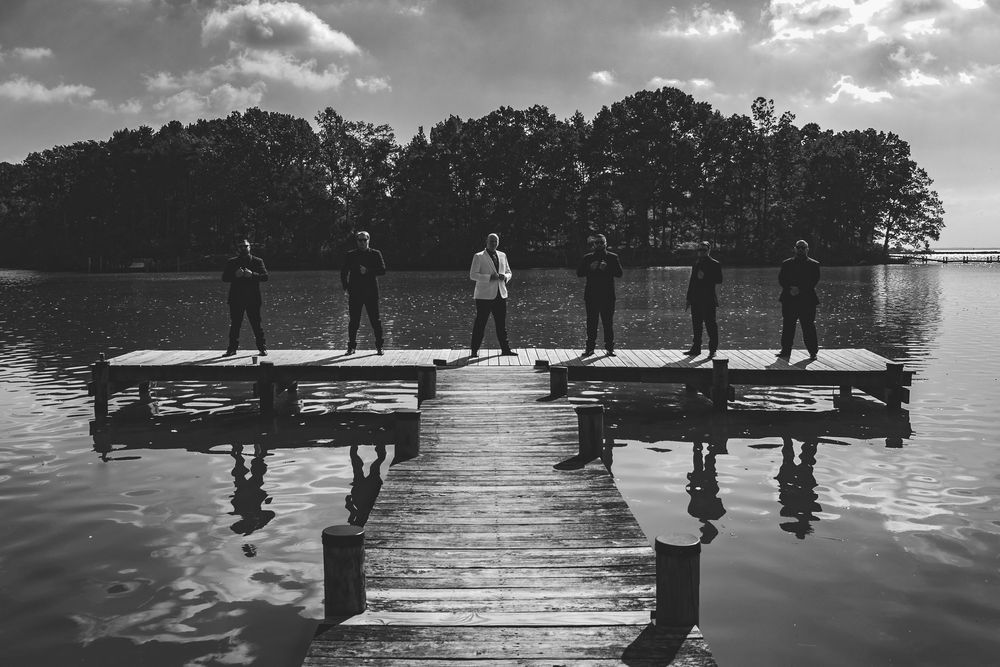 Silhouettes of people standing on a wooden dock over a lake with trees in the background on a cloudy day.