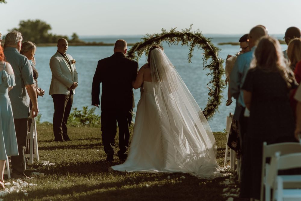 An outdoor wedding ceremony by a lake with guests gathered around a floral arch during golden hour.