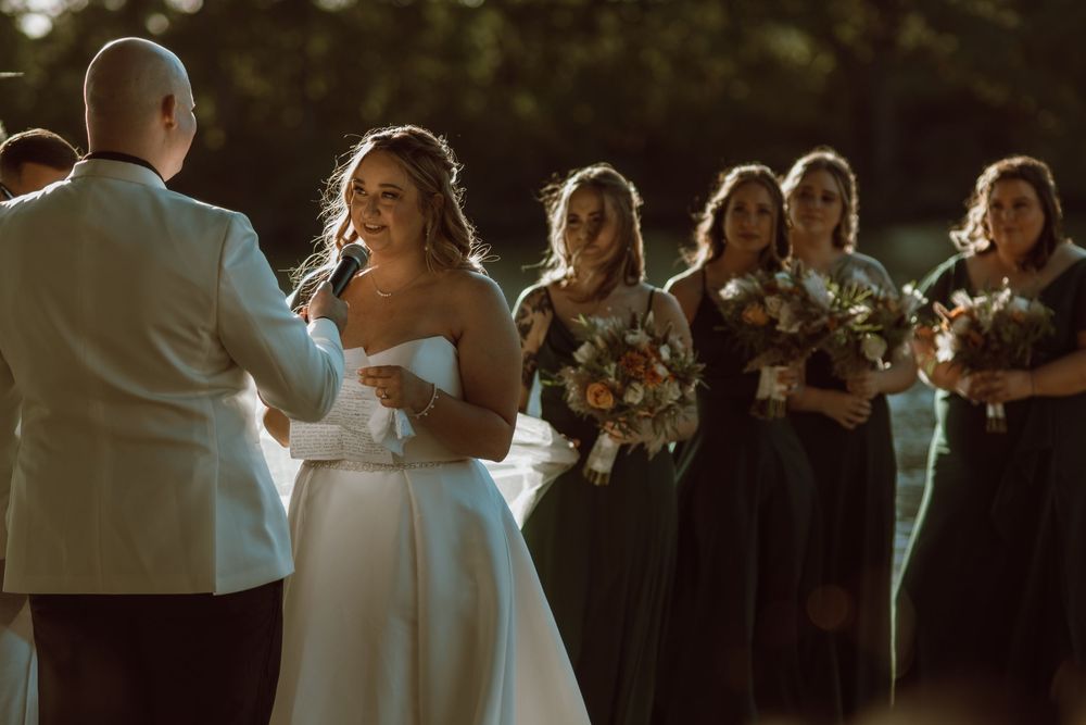A joyful wedding ceremony takes place outdoors at golden hour with bridesmaids in black dresses holding bouquets.