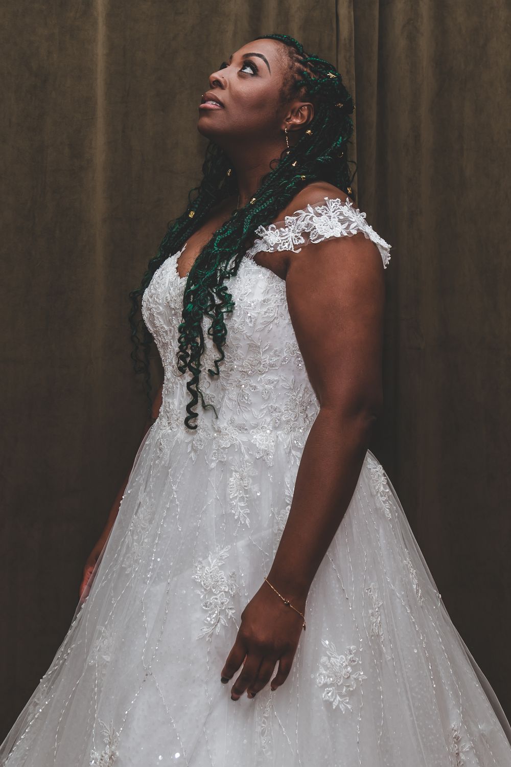 A bride in a detailed lace gown and braided hair gazes upward in a dramatic portrait.