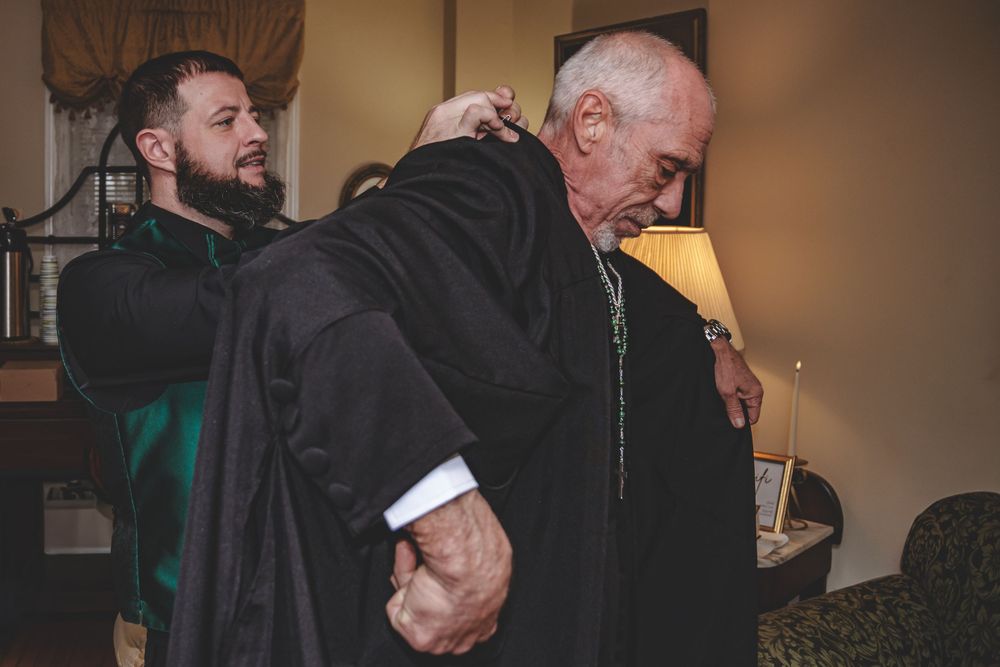 Two people help each other put on formal attire during what appears to be a wedding preparation moment.
