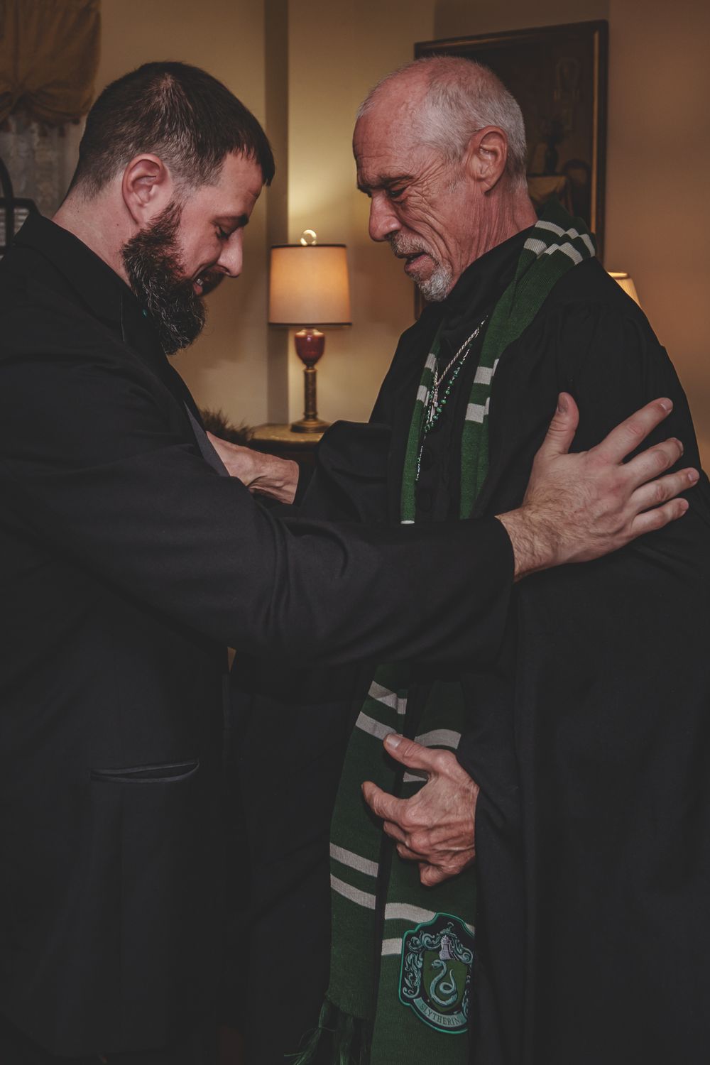 Two people share an intimate emotional moment in a dimly lit room, wearing a scarf with Celtic-style patterns.