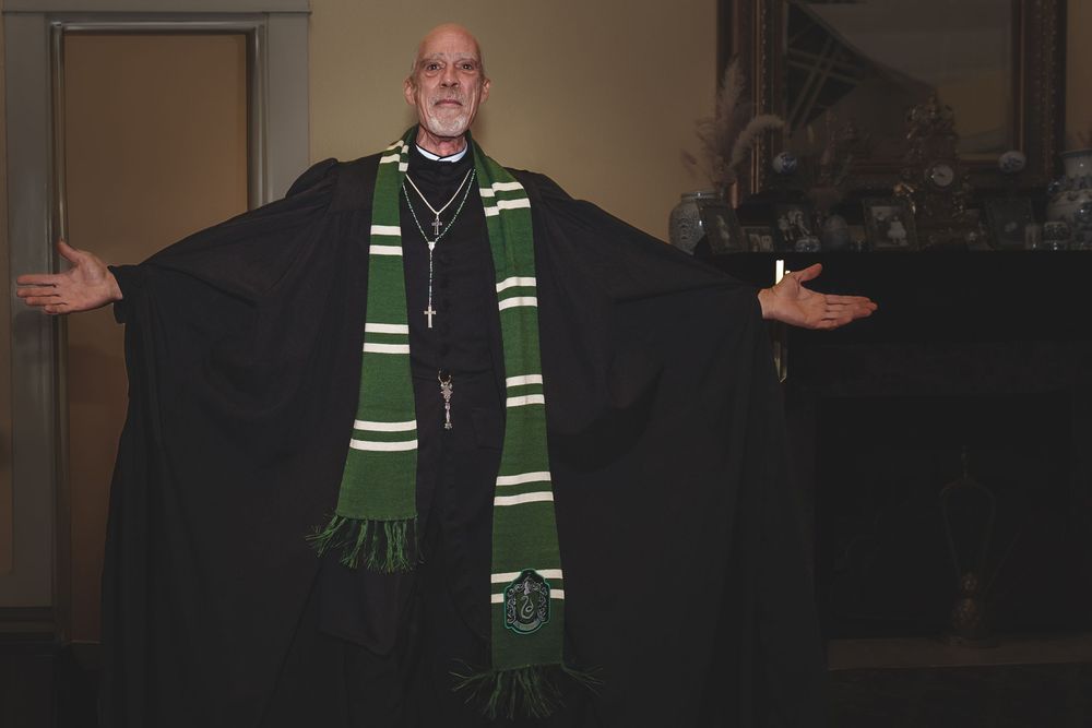 A person wearing a black robe and green striped scarf poses with outstretched arms.