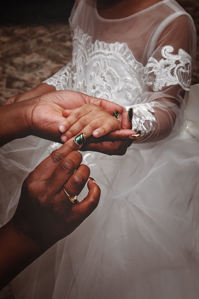 Close-up of hands exchanging rings against white lace wedding dress detail.