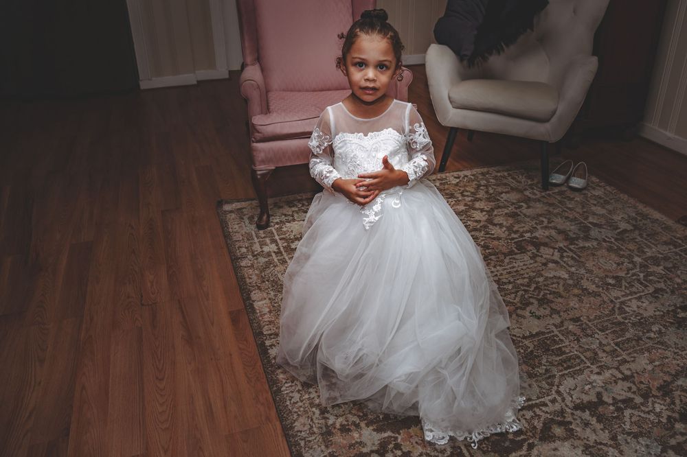 A young flower girl in a white tulle dress stands on a patterned carpet holding something in her hands.